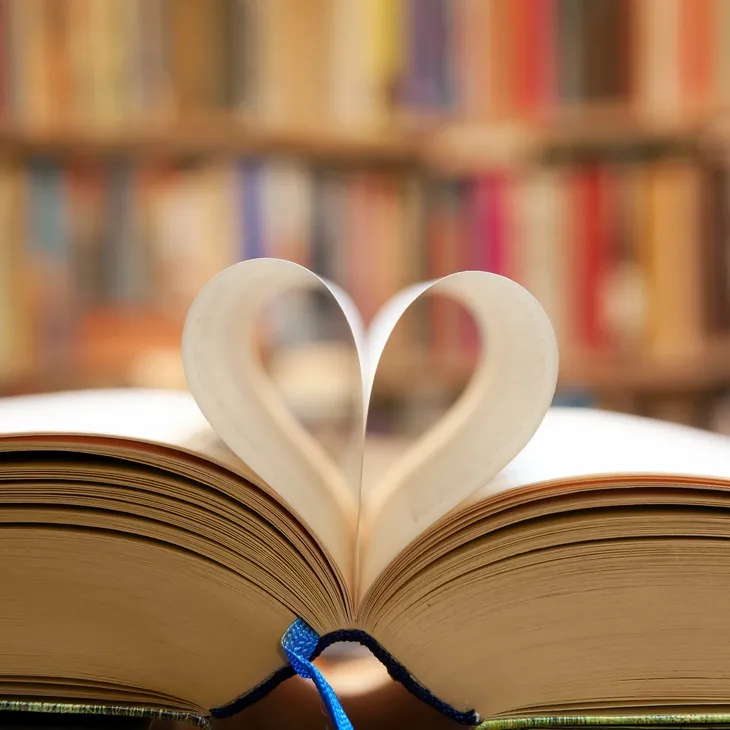 Open book with two pages curved inward to form a heart shape, symbolizing a Daily Affirmation, with blurred bookshelves in the background.